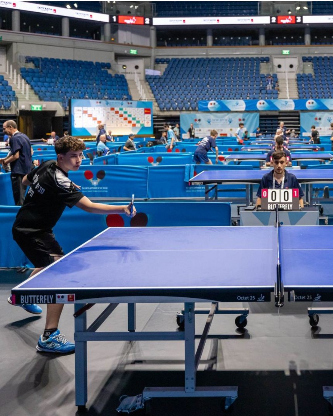 Dos jóvenes jugando un partido de tenis de mesa en un coliseo, con juez y marcador